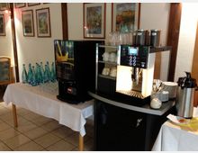 Tisch mit einer Kaffeemaschine, Milchkännchen und Tassen auf der rechten Seite sowie Wassergläsern und Wasserflaschen auf der linken Seite; im Hintergrund eine Wand mit gerahmten Gemälden.