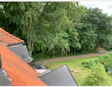 Dach mit orangen Ziegeln im Vordergrund, umgeben von dichtem Baumbestand. Im Hintergrund ein gepflasterter Weg, eine grasbewachsene Fläche mit Liegestühlen und ein Teich.