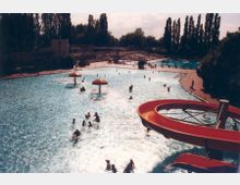 Ein Freibad mit mehreren Schwimmbecken, Wasserspielen und einer roten Wasserrutsche im Vordergrund. Im Hintergrund sind Liegeflächen, Bäume und ein Gebäude zu sehen.
