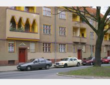 Mehrstöckiges Wohngebäude mit beiger Fassade und gelben sowie violetten Farbakzenten um Fenster und Balkone; markant sind gestufte Balkone im Vordergrund. Vor dem Gebäude parken mehrere Fahrzeuge, darunter ein Taxi, entlang einer Straße mit angrenzenden Bäumen.