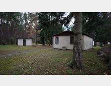 Zwei einfache, einstöckige Gebäude mit heller Fassade stehen in einem Waldgebiet, umgeben von Nadelbäumen und einer Rasenfläche. Im Vordergrund ist ein einzelner Baum sichtbar, und ein schmaler, gepflasterter Weg verläuft durch das Gelände.