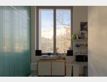 Heller Innenraum mit großem doppelflügeligem Fenster und Blick auf Wohngebäude und kahle Bäume. Unter dem Fenster stehen ein Sideboard mit Plattenspieler und Pflanzen sowie rechts offene Regale mit Geschirr und einer Mikrowelle; links eine halbtransparente grünliche Glaswand.