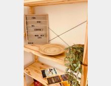 Holzregal an einer hellen Wand mit Querdrähten, darauf ein rundes Holztablett, Prospekte und eine hängende Grünpflanze. Links lehnt ein gerilltes Schild mit aufgedrucktem Text am Regal.