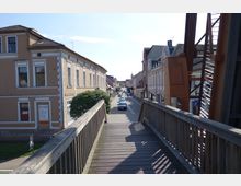 Holzbrücke, die zu einer Straße mit parkenden Autos und historischen Gebäuden auf beiden Seiten führt, darunter mehrstöckige Häuser mit verzierten Fensterrahmen und Schindeldächern in einer städtischen Umgebung. Rechts im Vordergrund ist ein Teil einer Stahlkonstruktion sichtbar.