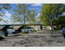 Flussufer mit gepflastertem Gehweg, Bänken und jungen Bäumen im Vordergrund; im Hintergrund eine breite Steinbrücke mit Bögen, darunter fließt ein ruhiger Fluss.