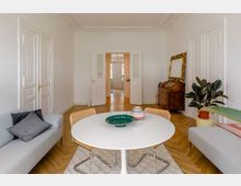 Helles Wohnzimmer mit Parkettboden, einem runden weißen Tisch und vier Stühlen, flankiert von zwei grauen Sofas. Im Hintergrund stehen eine Kommode, ein großer goldgerahmter Spiegel und eine Zimmerpflanze; doppelflügelige weiße Türen führen in einen weiteren Raum.