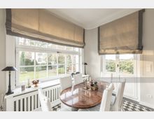 Esszimmer mit großen, weißen Sprossenfenstern, durch die ein Garten sichtbar ist. Der Raum ist mit einem runden Holztisch, weiß gepolsterten Stühlen und hellen, beigen Rollos ausgestattet; ein schwarz-weiß karierter Fliesenboden ergänzt das helle und elegante Design.