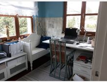 Ein kleiner Arbeitsplatz in einer hellen Ecke mit Holzrahmen-Fenstern. Ein weißer Schreibtisch mit Laptop und Schreibtischlampe steht vor den Fenstern, daneben stehen ein Regal mit Ordnern und eine Sitzbank.