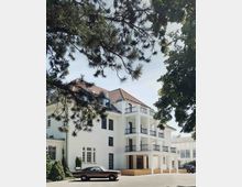 Ein weißes, dreistöckiges Gebäude mit einem roten Ziegeldach und Balkonen an der rechten Seite, umgeben von Bäumen. Im Vordergrund parken ein klassisches Auto und ein modernes Auto auf einer asphaltierten Fläche.