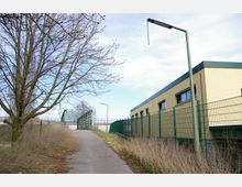 Gehweg entlang eines Zauns mit einer gelben Gebäudewand rechts, einem kahlen Baum links und einer Metallbrücke im Hintergrund. Die Umgebung ist von trockenen Sträuchern und vereinzelten Laternen geprägt.
