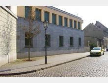 Straßenszene mit einem modernen Gebäude aus grauem Stein und gelber Fassade im oberen Bereich, kleinen rechteckigen Fenstern, einem Gehweg mit Kopfsteinpflaster sowie einem Laternenpfahl und einem jungen Baum. Im Vordergrund geparkte Autos auf einer gepflasterten Straße.