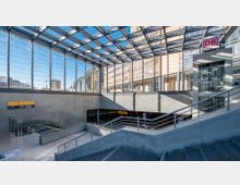 Modernes Bahnhofsgebäude mit einer großen verglasten Decke, durch die Tageslicht einfällt, und umliegenden Bürogebäuden. Mehrere Treppen und Rolltreppen führen zu einem unteren Bereich, wo Beschilderung und das Logo der Deutschen Bahn sichtbar sind.