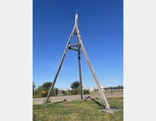 Holzstruktur in Form eines großen Dreibeins auf einer Wiese, gesichert mit Seilen. Im Hintergrund befindet sich ein asphaltierter Weg, Bäume und ein offenes Gelände, unter einem klaren, blauen Himmel.