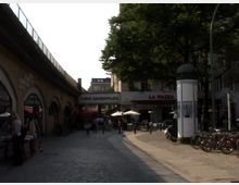 Straßenszene am Savignyplatz in Berlin mit einem erhöhten Bahngleis auf der linken Seite und einem kleinen Platz mit Cafétischen und Fahrrädern rechts. Im Hintergrund sind Geschäfte und Restaurants, darunter eines mit der Aufschrift "La Piazza", zu sehen.