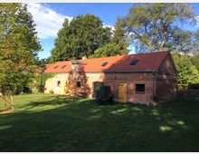 Langgestrecktes Backsteingebäude mit rotem Satteldach und mehreren Holztüren sowie kleinen Fenstern, umgeben von einer Wiese und Bäumen. Im Vordergrund steht ein Anhänger, und im Hintergrund ist ein Zaun erkennbar.