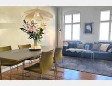 Wohnzimmer mit Essbereich in einem hellen Raum. Ein Esstisch mit grünen Stühlen und einem Blumenarrangement steht auf einem Holzboden, während im Hintergrund ein Sofa mit Kissen und einer Decke vor drei großen Fenstern zu sehen ist.