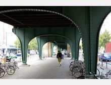 Eine Unterführung mit einer grünen Stahlkonstruktion, die von genieteten Stützpfeilern getragen wird. Unter der Brücke stehen geparkte Fahrräder, und Menschen gehen auf einem gepflasterten Weg, während im Hintergrund eine städtische Straße mit Autos und Geschäften sichtbar ist.