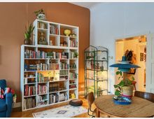 Ein Wohnzimmer mit einer weißen Regalwand vor einer orangefarbenen Wand, gefüllt mit Büchern, Pflanzen und Dekorationsgegenständen. Im Vordergrund steht ein runder Holztisch mit einer blauen Hängelampe und einer Pflanze in einem Korb; rechts ist ein kleiner Pflanzenvorratsständer aus Glas und Metall zu sehen.