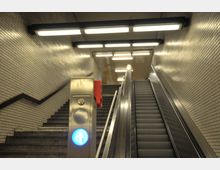 Treppenaufgang und Rolltreppe in einer U-Bahn-Station mit Wänden aus weißen Kacheln. Im Vordergrund ein Edelstahlpfosten mit einem leuchtenden blauen Pfeil nach oben.