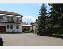 Zweistöckiges weißes Wohnhaus mit braunem Ziegeldach und rotem Geländer am Balkon. Vor dem Haus befinden sich ein gepflasterter Hof, eine Sitzbank und ein großer Tannenbaum; im Hintergrund stehen weitere Gebäude.