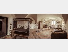 Historischer Innenraum mit Gewölbedecke, rustikalem Mobiliar und Steinwänden. Links ein Himmelbett und ein Holztisch, rechts ein Sitzbereich mit Sofa, ein Holztisch sowie ein Esstisch vor einem Fenster mit sichtbaren Holzbalken.