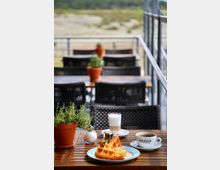 Ein Außenbereich eines Cafés mit Holztischen und schwarzen Flechtstühlen; auf einem vorderen Tisch stehen eine Tasse Kaffee, ein Glas Latte Macchiato, eine Portion Waffeln mit Soße und Kräuterpflanzen in Tontöpfen. Im Hintergrund ist eine Landschaft mit Wiese und Geländer zu sehen.