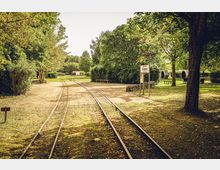 Schmalspurbahn-Gleise in einem Park mit umliegenden Bäumen und Bänken; im Hintergrund ein Schild mit der Aufschrift „La Sylviane“ und moderne Outdoor-Sitzmodule.