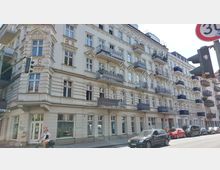 Mehrstöckiges Altbau-Wohnhaus mit verzierten Fassaden, klassischen Stuckelementen und Balkonen an fast jeder Wohnung, gelegen an einer Straßenecke mit parkenden Autos und einer Fahrradfahrerin im Vordergrund. Ein Straßenschild und eine Ampel sind am rechten Bildrand zu sehen.