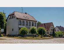 Ein kleines Postamtgebäude in heller Fassade, versehen mit roten Dachziegeln und großen Fenstern, steht an einer gepflasterten Straße. Daneben befinden sich zwei ähnliche Wohnhäuser mit grünen Bäumen und Gärten im Vordergrund.