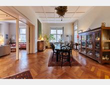 Wohnzimmer und Essbereich in einem klassisch eingerichteten Altbau mit Holzboden im Fischgrätmuster. Zu sehen sind ein Esstisch mit schwarzen Stühlen, ein antikes Holzbuffet, große Fenster mit Blick auf Bäume sowie ein angrenzender Wohnbereich mit Polstermöbeln.