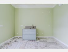 Leerer Küchenraum mit hellgrünen Wänden, Stuckdecke und marmoriertem Steinboden. In der Mitte steht ein grauer Unterschrank mit Spüle und Armatur; mehrere Steckdosen und Anschlüsse sind an der Wand verteilt.