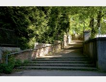 Fußweg mit leichtem Anstieg, gesäumt von einer Ziegelmauer und dichter Hecke auf der linken Seite. Auf der rechten Seite befindet sich eine Betonmauer, beides ergänzt durch Geländer, mit Bäumen und Sonnenschein im Hintergrund.