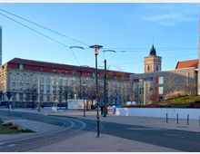 Straßenszene mit einem historischen Gebäude mit roten Ziegeldach und zahlreichen Fenstern im Hintergrund, daneben ein moderner Bau aus grauen Backsteinen. Im Vordergrund verlaufen Straßenbahnschienen, und Laternenmasten heben sich gegen den blauen Himmel ab.