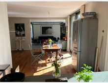 Moderne Wohnküche mit Holzboden, einem Holztisch mit Metallstühlen, einer Edelstahl-Kühl-Gefrierkombination und großen Fenstern mit Vorhängen. Im Hintergrund ist ein offenes Wohnzimmer mit Sofa, Wandkunst und weiteren Einrichtungsgegenständen sichtbar.