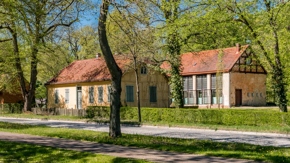 Low, historic buildings with faded stucco walls and red tile roofs sit along a quiet road in a leafy park setting. Tall trees surround the structures, including one section with large multi-pane windows and timber framing on the gable.