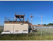 Flaches Gebäude mit einer Außentreppe und einem Plattformaufbau, umgeben von einem Metallzaun. Im Hintergrund sind zwei rote Funkmasten auf einer Wiese zu sehen, vor einem klaren blauen Himmel.