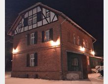 Backsteingebäude im Fachwerkstil mit beleuchteten Fenstern und weißem Schnee im Vordergrund, aufgenommen bei Nacht. Das obere Geschoss zeigt ein Fachwerkdesign, während die unteren Fassaden aus unverputztem Ziegelstein bestehen.