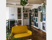 Ein heller Innenraum mit Holzdielenboden, einer gelben Couch im Vordergrund und mehreren Pflanzen. An der Wand hängen zahlreiche gerahmte Schallplatten und Bilder; im Hintergrund ist durch verglaste Trennwände ein Bürobereich erkennbar.