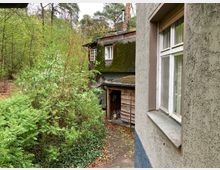 Außenansicht einer verwitterten Hausfassade, umgeben von dichtem Grün und Bäumen, mit einem Nebengebäude im Hintergrund, dessen Dach mit Moos bewachsen ist. Der Boden ist von Laub bedeckt, und die Szene liegt in einem bewaldeten Gebiet.