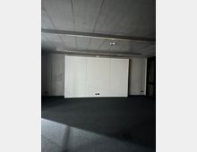 Ein leerer Raum mit Betonwänden und -decke, einer weißen Wandverkleidung in der Mitte und einem dunklen Teppichboden. An der Decke sind schmale, lange Leuchtleisten angebracht, und an der weißen Wand befinden sich zwei schwarze Steckdosen.