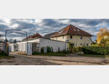 Eingangsbereich einer kleinen Freizeitanlage mit dem Schriftzug "SG Wasserwandern" über einem Metalltor. Im Hintergrund stehen ein beiges Wohnhaus mit Ziegeldach und eine Reihe von weißen Garagen; umgeben von Büschen und herbstlicher Vegetation.