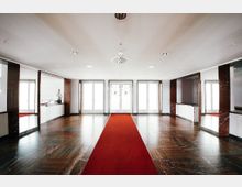 Großer, heller Eingangsbereich mit Hochglanz-Holzboden und rotem Teppich, der zu drei doppelflügeligen Glastüren führt. Die Wände sind teilweise mit dunklem Marmor verkleidet, und die Decke ist mit eingelassenen Spotlichtern und Belüftungselementen versehen.