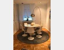 Esszimmer mit rundem Marmortisch und sechs weißen Schalenstühlen im Retro-Stil auf einem runden Teppich. Im Hintergrund steht vor einem Fenster mit weißen Vorhängen eine Vase mit getrockneten Gräsern auf einer Heizung, darüber eine gewölbte Stehleuchte mit silbernem Schirm.