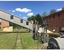 Industrielle Außenanlage mit einer alten Schienenstrecke, einem Förderband aus Metall mit Fensterreihen, mehreren gemauerten und metallenen Bauten sowie einem roten Backsteingebäude im Hintergrund. Die Umgebung ist von Grasflächen und vereinzelten Bäumen geprägt unter einem blauen Himmel mit einzelnen Wolken.