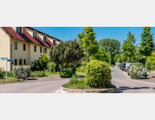 Wohnstraße in einem Vorort mit einem gelben Reihenhaus und rotem Dach links im Bild. Die Straße ist von Bäumen, Sträuchern und geparkten Autos gesäumt, mit einem Kreisverkehr im Vordergrund und einem klaren blauen Himmel im Hintergrund.