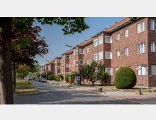 Straßenansicht mit Reihen von Backsteingebäuden im Stil des 20. Jahrhunderts, charakterisiert durch weiße Fensterrahmen und mehrere Etagen. Im Vordergrund befinden sich ein Gehweg, eine Bushaltestelle mit wartenden Personen und gepflegte Bäume sowie geometrisch geschnittene Büsche.