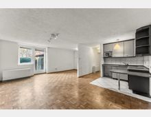 Heller, leerer Wohnraum mit Parkettboden und weißen Wänden, mit großem Fenster und Glastür zum Balkon. Rechts befindet sich eine offene Küchenzeile mit grauen Schränken, gefliester Rückwand und kleinem Tresen auf einem gefliesten Bodenbereich.