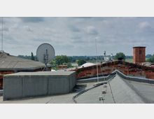 Ansicht eines Ziegeldachs mit diversen technischen Elementen wie einer Satellitenschüssel und Kabeln, im Hintergrund eine Landschaft mit Bäumen, einem Fluss und einer Brücke. Daneben steht ein roter Backsteinturm.