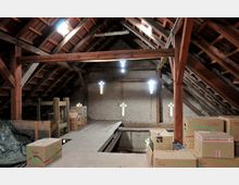 Dachboden in einem alten Gebäude mit sichtbarer Holzkonstruktion und Ziegeln im Dach. Der Raum enthält mehrere Pappkartons und Holzbalken, die Tageslicht fällt durch kreuzförmige Fensteröffnungen in der Rückwand.