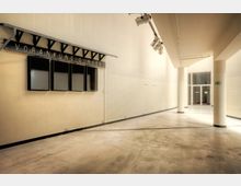 Weitläufiger, heller Innenraum mit glatten Betonböden und schlichten, beigefarbenen Wänden. Links hängen leere schwarze Anzeigetafeln mit der Aufschrift „Vorankündigungen“, rechts befinden sich helle Säulen und ein Fluchtwegzeichen; im Hintergrund ist eine Glastür erkennbar.